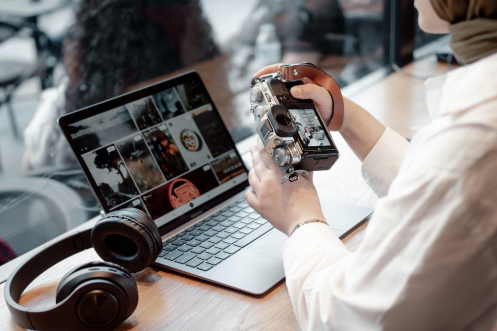 A woman editing photos on her laptop while holding a camera, with headphones nearby on a desk.